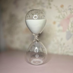 May include: A clear glass hourglass with white sand inside. The top and bottom bulbs are decorated with etched floral designs. The hourglass is set against a soft pink surface and a floral wallpaper background.