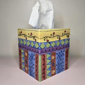May include: A decorative tissue box with a floral and geometric pattern. The top section is yellow with a vine of red flowers. The middle section has a diamond pattern in green and purple. The bottom section has vertical stripes in blue, red, and green.