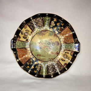 May include: A decorative ceramic plate with a detailed bird and floral design. The plate features a gold center with a bird and landscape scene, surrounded by a patterned border in black, gold, green, and red. The plate has a scalloped edge.