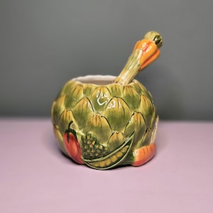 May include: A ceramic artichoke-shaped container with a matching spoon. The container is green and yellow with painted vegetables, including a tomato, peas, and carrots. The spoon handle is shaped like a carrot and pepper.