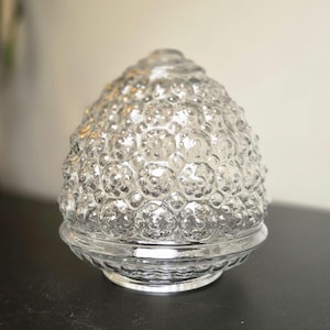 May include: A clear glass light fixture shade with a rounded, dome-like shape. The surface features a repeating, raised hobnail or bubble texture throughout the exterior, ending in a smooth, circular base rim designed for mounting to a light fixture.