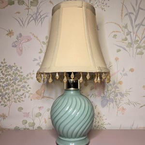 May include: A table lamp with a light blue, spirally ribbed ceramic base and a metallic silver neck. The lamp features a cream-colored fabric shade with a straight-sided design, accented by a fringe of clear, teardrop-shaped glass beads.