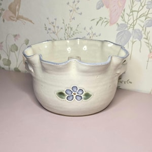 May include: A cream-colored ceramic bowl with a ruffled rim and a light blue trim. The bowl features a hand-painted blue flower with green leaves on the front. The bowl has two small handles and a hole in the center.