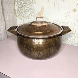 May include: A vintage copper pot with a lid. The pot has a hammered texture and a tarnished finish, with a silver rim and handle. The lid has a small handle. The pot has two square handles on either side.