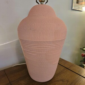 May include: A vintage pink table lamp with a textured surface. The lamp has a bulbous top and a flared base. The lamp's metal hardware is brass-coloured. The lamp is sitting on a wooden table.