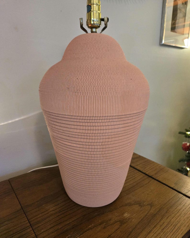 May include: A vintage pink table lamp with a textured surface. The lamp has a bulbous top and a flared base. The lamp's metal hardware is brass-coloured. The lamp is sitting on a wooden table.