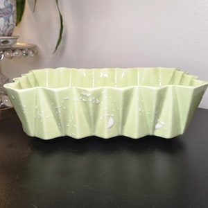 May include: A rectangular ceramic dish with a light green glazed finish and a pleated, accordion-style side design. The exterior surface features a subtle, white speckled pattern distributed across the vertical folds of the vessel.
