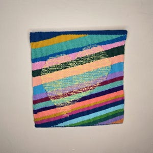 May include: A colorful woven wall hanging with a geometric design. The artwork features diagonal stripes in shades of blue, green, pink, orange, and yellow. The woven texture is visible, adding depth and visual interest to the piece.