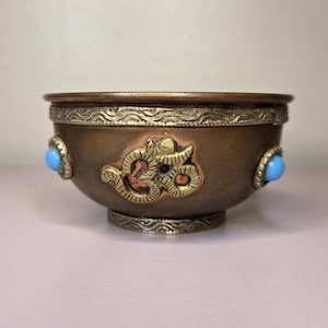 May include: A bronze-colored bowl with ornate gold-colored detailing and two light blue stones. The bowl features a decorative design in gold and is likely used for ceremonial purposes.