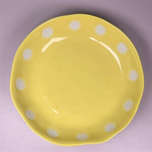 May include: A yellow ceramic plate with a wavy edge. The plate is decorated with white starburst patterns around the rim. The plate has a glossy finish and is set against a light purple background.