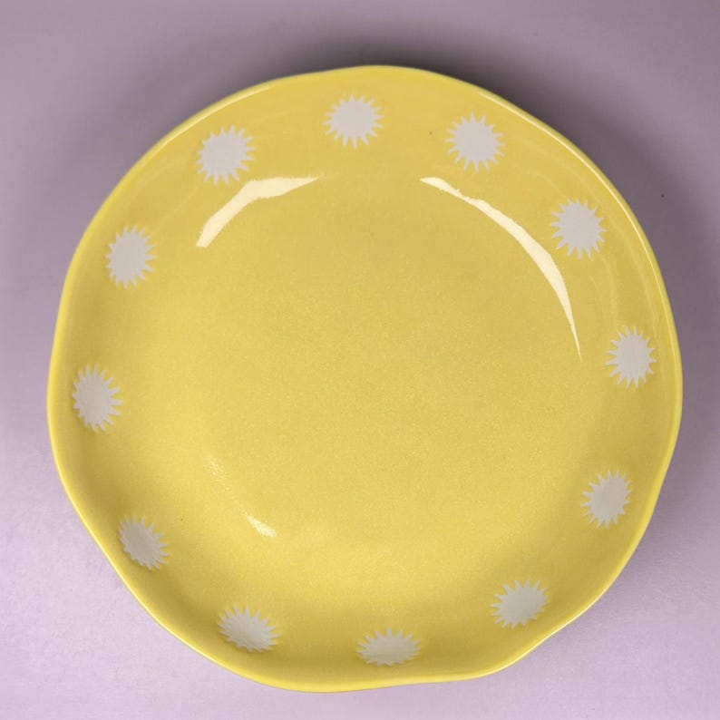 May include: A yellow ceramic plate with a wavy edge. The plate is decorated with white starburst patterns around the rim. The plate has a glossy finish and is set against a light purple background.
