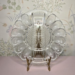 Puede incluir: Una fuente de cristal transparente para servir ostras con un soporte decorativo de metal dorado. La fuente tiene un compartimento central y varias secciones con forma de ostra alrededor del borde. El cristal tiene un patrón texturizado.