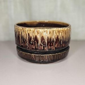 May include: A two-piece ceramic planter with a brown and tan glaze. The top section is a bowl shape, and the bottom section is a matching saucer. The glaze has a dripped effect, creating a unique texture. The planter is suitable for indoor plants.