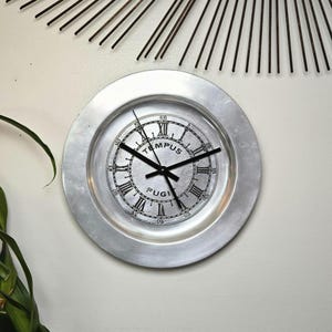 May include: A circular wall clock with a wide, brushed silver-tone metal frame. The clock face features black Roman numerals, black hands, and the text "TEMPUS FUGIT" printed in black. The design includes a secondary minute track with numbers.