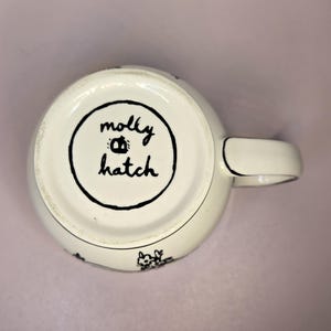 May include: A white ceramic mug with a black handle and the text "But First, Coffee" in a bold, black font. The mug is set against a blurred background, suggesting a cozy setting.