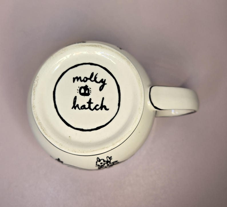 May include: A white ceramic mug with a black handle and the text "But First, Coffee" in a bold, black font. The mug is set against a blurred background, suggesting a cozy setting.