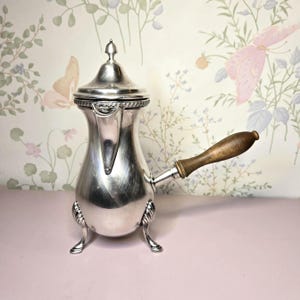 May include: A silver-colored coffee pot with a wooden handle. The pot has a lid with a small finial and sits on three decorative feet. The handle is attached to the side of the pot. The background features a floral wallpaper.