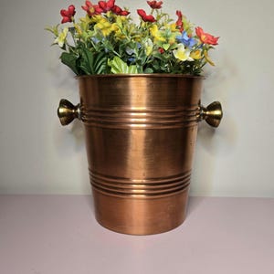 May include: A copper-colored metal bucket with two decorative handles, filled with colorful artificial flowers. The bucket has horizontal bands around its body. The flowers include red, yellow, and blue blossoms, creating a vibrant display.