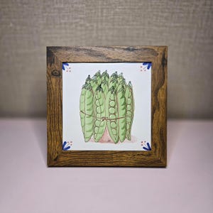 May include: Square wooden framed art featuring a watercolor illustration of a bundle of green pea pods tied with a brown string. The artwork is on a white tile with blue and pink floral accents in the corners.