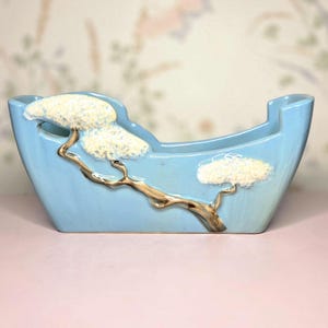 May include: A light blue ceramic planter with a boat-like shape. The planter features a brown tree branch design with white, textured tree tops. The planter is designed for decorative use.