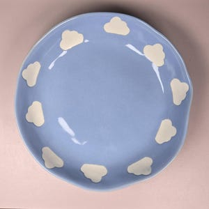May include: A light blue ceramic plate with a wavy edge. The plate is decorated with cream-colored cloud shapes around the rim. The plate is round and appears to be handmade.