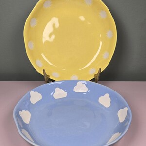 May include: Two ceramic bowls. The top bowl is yellow with white starburst designs around the rim. The bottom bowl is light blue with white cloud designs around the rim. Both bowls have a glossy finish.