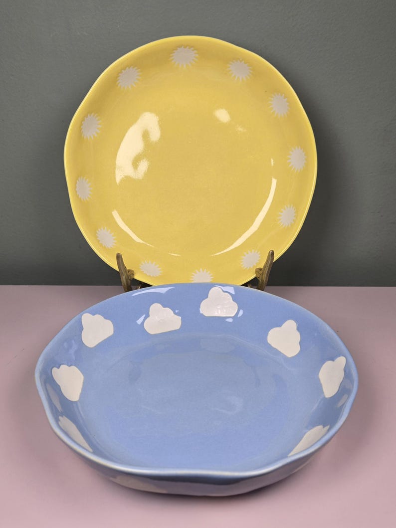 May include: Two ceramic bowls. The top bowl is yellow with white starburst designs around the rim. The bottom bowl is light blue with white cloud designs around the rim. Both bowls have a glossy finish.