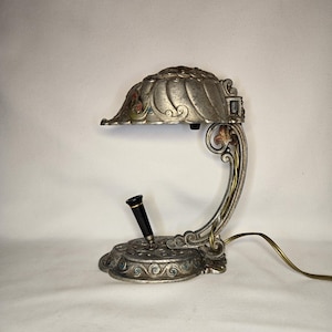 May include: An antique desk lamp with a silver finish and ornate floral detailing. The lamp features a curved arm, a domed shade, and a pen holder. The shade has red, green, and yellow accents. A black pen is in the holder.