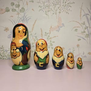 May include: A set of five wooden nesting dolls, featuring Snow White and the Seven Dwarfs. The largest doll depicts Snow White, while the smaller dolls represent the dwarfs. Each doll is painted with vibrant colors and detailed facial expressions.