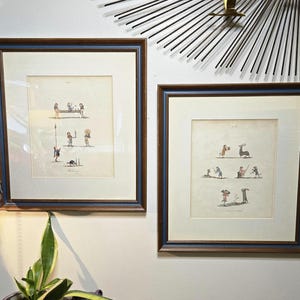 May include: Two framed antique prints with blue and brown frames. The prints feature small, detailed illustrations of figures in various scenes. The artwork is displayed on a light-colored wall, with a decorative lamp and plant in the foreground.