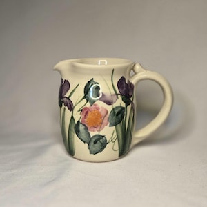 May include: Cream-colored ceramic pitcher with a floral design. The pitcher features purple irises, a pink flower, and green leaves. It has a curved handle and a spout for pouring. The flowers are painted on the front of the pitcher.
