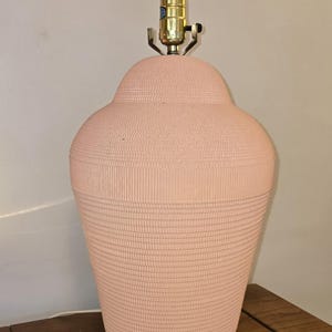 May include: A peach-coloured ceramic table lamp with a textured surface. The lamp has a bulb and brass-coloured hardware. The lamp is on a wooden table. The lamp's design is a classic, decorative style.