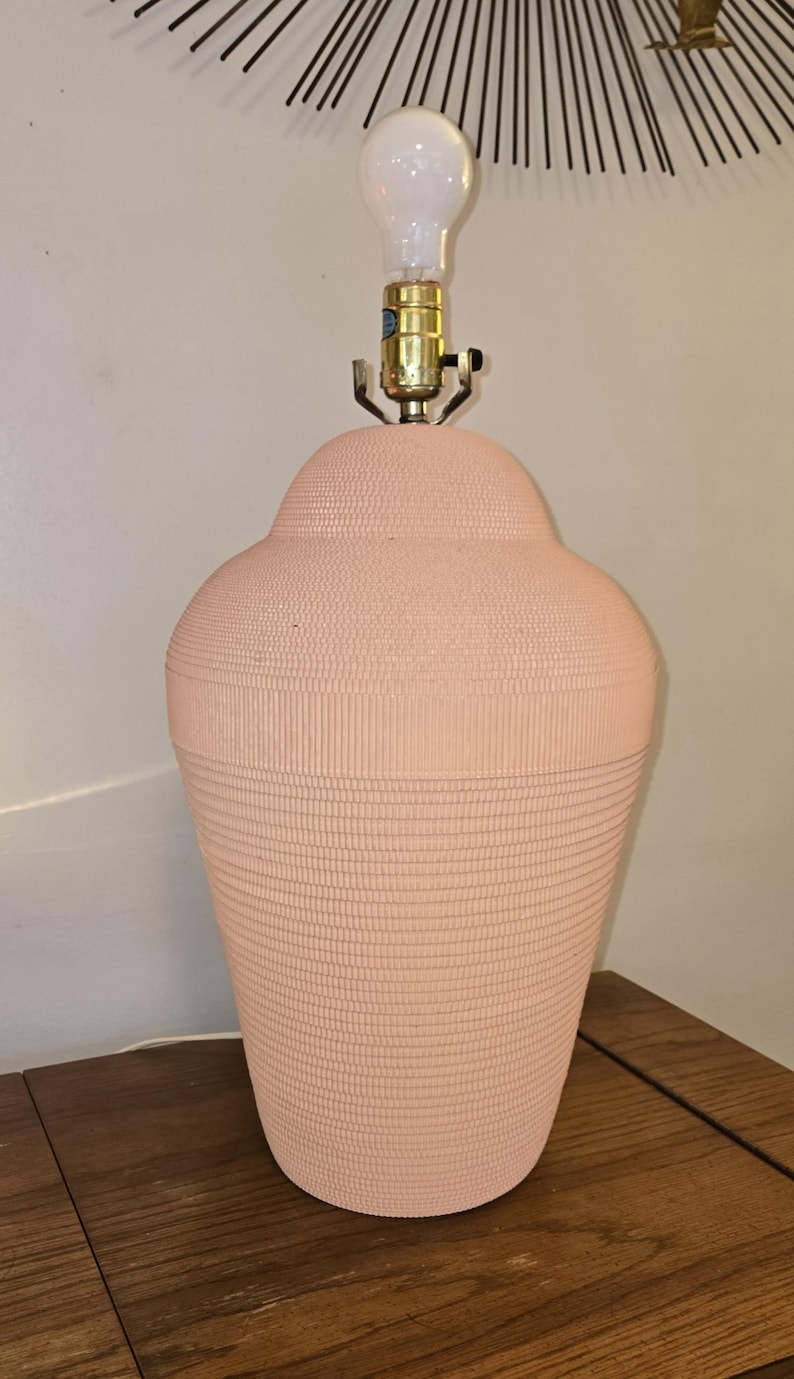 May include: A peach-coloured ceramic table lamp with a textured surface. The lamp has a bulb and brass-coloured hardware. The lamp is on a wooden table. The lamp's design is a classic, decorative style.