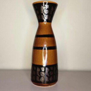 May include: A tall ceramic vase with a flared rim, featuring a glossy brown glaze accented by horizontal black bands and decorative black panels at the top and bottom that display a repeating, textured, organic pattern in a dark finish.