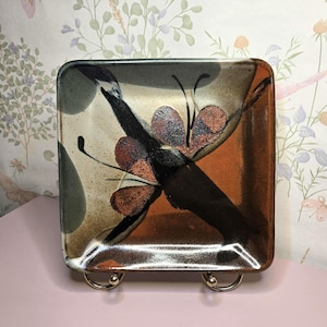 May include: Square ceramic plate with a butterfly design. The plate features a brown, orange, and cream color scheme with a dark brown butterfly design. The plate is displayed on a metal stand.