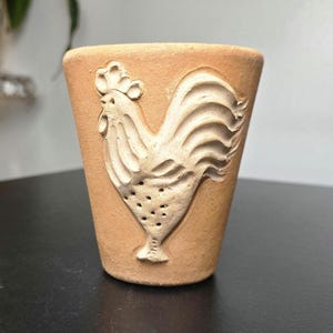 May include: A light brown terracotta cup featuring a raised, embossed relief of a rooster on the front surface. The cup has a tapered, conical shape with a smooth rim and a matte, unglazed finish that highlights the textured clay material.