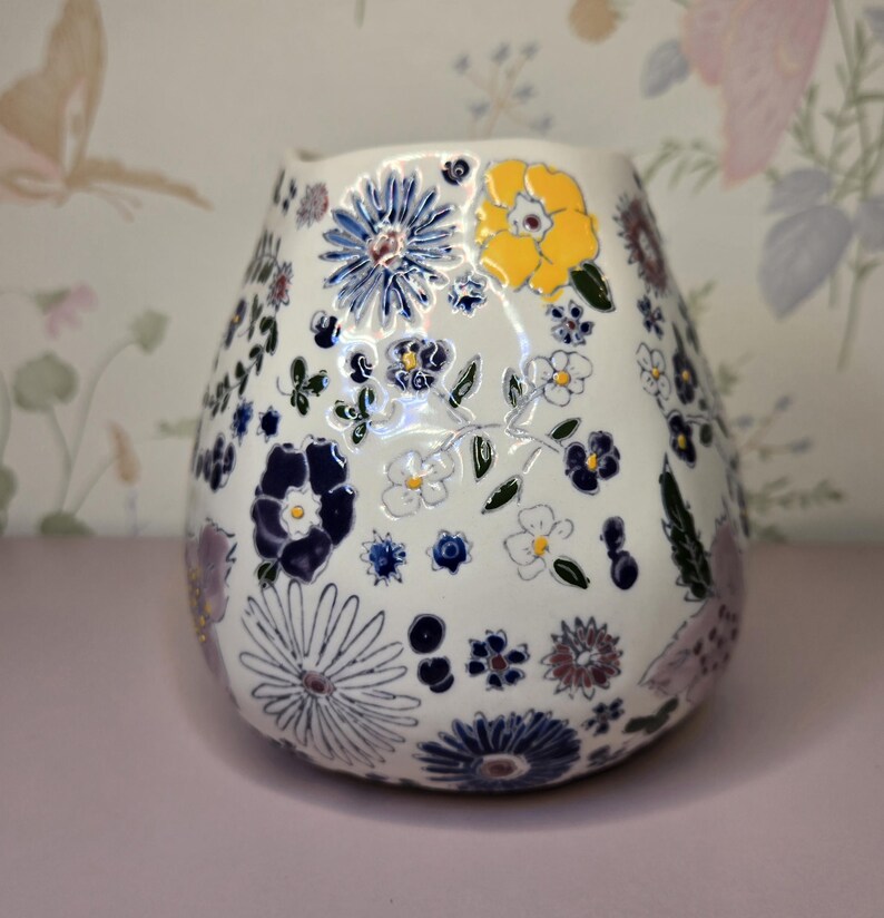 May include: White ceramic vase with a floral pattern. The vase is decorated with blue, yellow, and purple flowers, along with green leaves. The vase has a rounded shape and a slightly flared top.