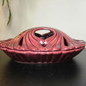 May include: A glossy, deep plum-colored ceramic pie dish with a matching lid. The lid features a decorative, radiating ribbed pattern and a central arched handle with integrated ventilation cutouts, creating a distinct, symmetrical design.