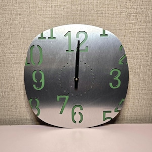 May include: A round, brushed metal wall clock with green numbers and hands. The numbers are cut out, revealing a green backing. The clock has a simple, modern design, perfect for any room. The clock hands point to 12 o'clock.