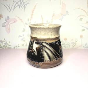 May include: A ceramic candle holder with a star and comet design. The holder is a mix of brown, black, and cream colors. The star and comet shapes are cut out, allowing light to shine through. The top rim is cream-colored.