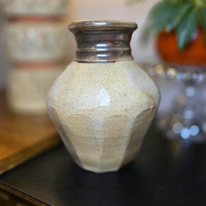 May include: A handcrafted ceramic vase with a light beige body and a dark brown rim. The vase has a textured surface and a unique, faceted design. The vase is a decorative item for the home.
