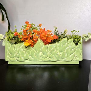 May include: A rectangular light green ceramic planter featuring a raised relief pattern of stylized leaves on its exterior. The planter is filled with a vibrant arrangement of bright orange and light green artificial flowers and foliage.