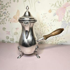 May include: A silver-toned metal chocolate pot with a bulbous body, a pointed lid, and three ornate feet. The vessel features a long, straight wooden handle extending from the side and a small spout with a decorative band below the lid.
