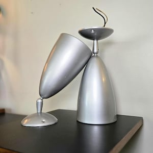 May include: Two silver-colored metal wall lamps with conical shades. Each lamp features a circular base, a short adjustable arm with a pivot joint, and a smooth metallic finish. One lamp is mounted upright, while the other is tilted at an angle.