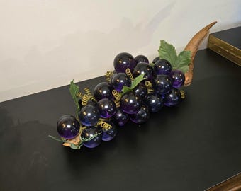 May include: A decorative cluster of translucent, deep purple glass grapes attached to a piece of natural driftwood. The arrangement includes green fabric leaves and thin, coiled gold-coloured wire tendrils interspersed among the round glass spheres.