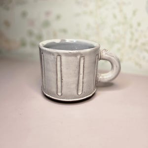 May include: A handmade ceramic mug in a light gray color with vertical, recessed lines. The mug has a rounded handle and a dark base. The interior is a darker gray. The mug is a unique piece of pottery.