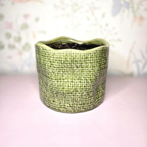 May include: A small, green ceramic planter with a textured, woven-like pattern. The rim has a wavy design. The interior is dark, likely filled with soil. The planter sits on a pink surface, with a floral patterned background.