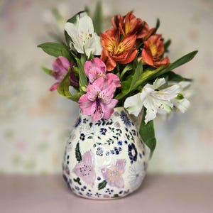May include: A white ceramic vase with a rounded shape, decorated with a hand-painted pattern of small blue, purple, and green flowers. The vase is filled with a bouquet of fresh alstroemeria flowers in shades of white, pink, and orange.