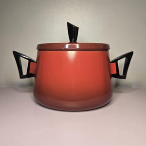 May include: A red enamel pot with black handles and a black lid. The pot has a rounded shape and a slightly flared rim.