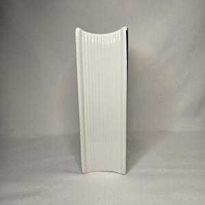May include: A white ceramic object with a vertical ribbed texture and a concave top and bottom edge. The object has a rectangular profile with rounded corners and a smooth, glossy finish, standing upright on its side to display the ribbed surface.